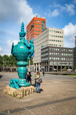 Blauwgroen keramisch kunstwerk op het Johanna Westerdijkplein in Den Haag met moderne kantoorgebouwen op de achtergrond.